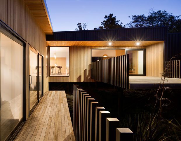 Twilight view of a contemporary courtyard-like residential exterior with warm timber-lined soffits and walls, extensive glazing, timber decking, and dark vertical slatted screening and fencing. Interior spaces are visible through the windows, with integrated warm lighting emphasizing the layered outdoor circulation and private courtyard design.