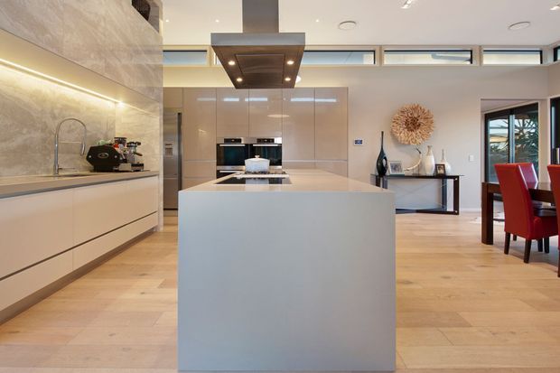 Contemporary open-plan kitchen featuring a large minimalist island with a pale stone benchtop and waterfall end, glossy light cabinetry, integrated appliances, a sculptural ceiling-mounted rangehood, and warm light timber flooring. A dining area with red upholstered chairs is visible to the right, while a textured stone backsplash and under-cabinet lighting add visual interest.