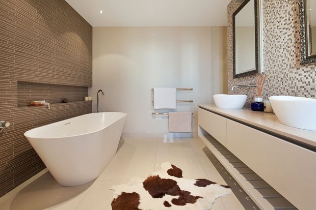 A spacious contemporary bathroom featuring a freestanding white bathtub, double vessel basins on a floating vanity, a large mirrored mosaic-tile feature wall, and a textured horizontal wall finish behind the bath. Pale tiled flooring and integrated towel rails create a clean, refined interior.
