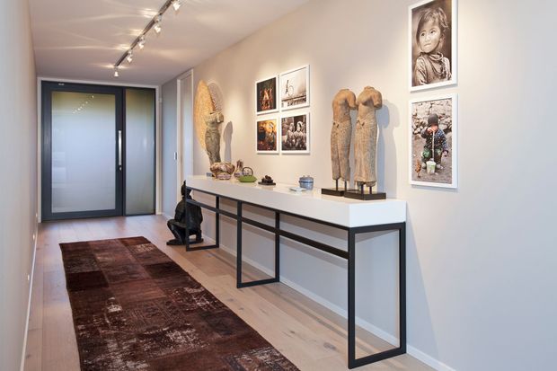 A refined residential hallway or entry gallery with pale walls and light timber flooring. A long, slim black-framed console table with a white top runs along the wall, styled with sculptural objects and artwork. Framed photographic and artistic pieces create a gallery-like display, while a dark patterned runner adds contrast. Black-framed glazed doors and ceiling-mounted track lighting extend toward the rear.