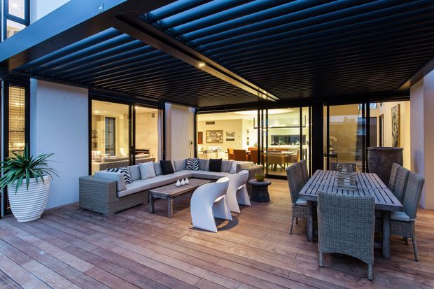 A covered outdoor entertaining area with timber decking, a dark slatted pergola ceiling, and extensive glass walls opening to a bright interior. The space includes a lounge setting with sofas and armchairs, plus a large outdoor dining table with woven chairs. Planters and warm integrated lighting add a relaxed, contemporary atmosphere.