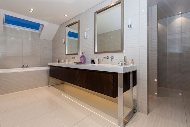Contemporary bathroom with a freestanding bathtub, wide double vanity, dark timber cabinetry, white benchtop, large framed mirrors, pale tiled walls and floor, and a glass-enclosed shower. A skylight and recessed lighting create a bright, refined atmosphere.