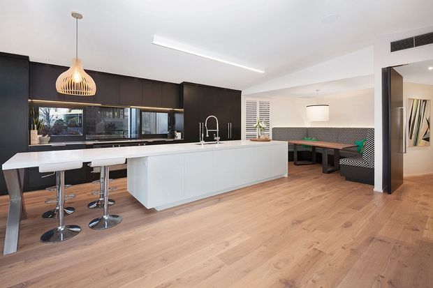 Open-plan contemporary kitchen and dining area with a large white island and breakfast bar, dark full-height cabinetry, integrated appliances, timber flooring, pendant lighting, and a built-in dining bench with patterned upholstery. The space is bright and spacious with a clean, refined finish.