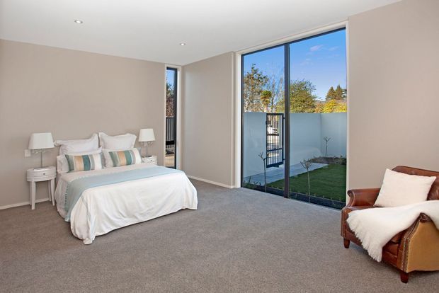 Bright, spacious contemporary bedroom with a neatly made bed, bedside tables and lamps, neutral painted walls, grey carpet, and full-height dark-framed glazing opening views toward a small landscaped courtyard. A leather armchair with a light throw sits in the corner.