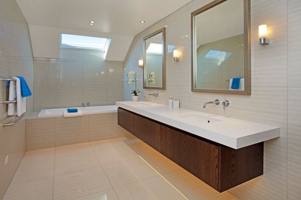 A bright, contemporary bathroom with full-height beige tiled walls and matching floor tiles. The room features a long floating dark timber vanity with a white stone benchtop, twin basins, wall-mounted chrome taps, large framed mirrors, recessed ceiling lights, a skylight, a built-in bathtub, and warm wall sconces.