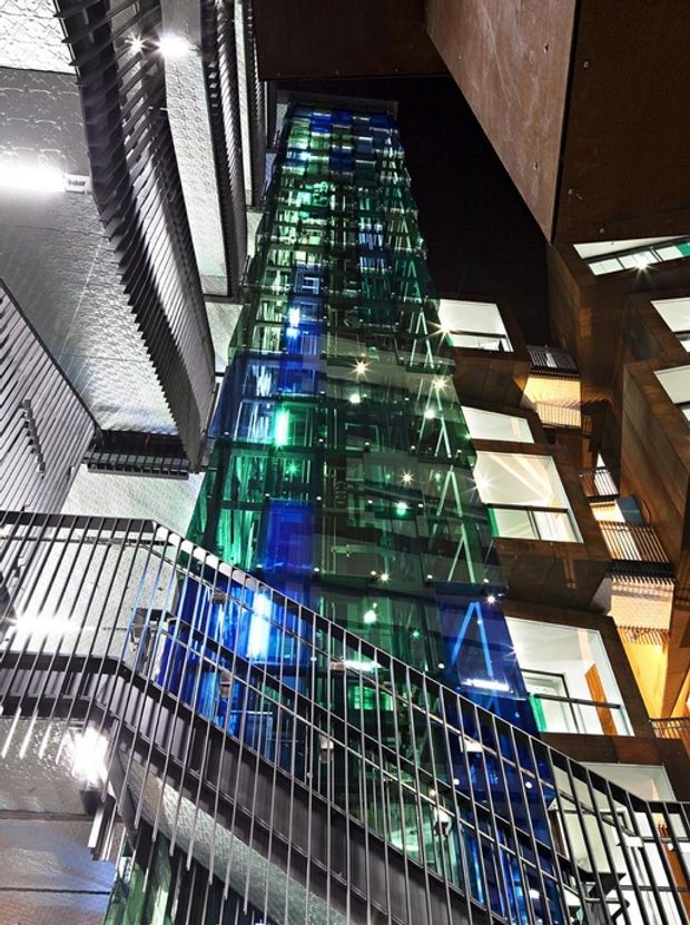 Upward-looking nighttime view of a dense modern multi-storey building courtyard or atrium. A tall illuminated glass elevator or glazed vertical circulation shaft dominates the centre, glowing in blue and green, surrounded by dark metal louvres, exposed balconies, railings, warm timber-toned cladding and large windows. Strong architectural lighting and dramatic perspective create an urban commercial or mixed-use character.