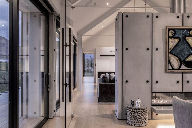 A refined contemporary interior viewed from a glazed entry or circulation area, featuring full-height black-framed glazing, pale polished flooring, white walls and a high sloped ceiling. A dramatic concrete-panel partition with exposed dark fasteners forms a fireplace feature and separates the corridor from a living area. A dark sofa, sculptural side table, artwork and partial upholstered seating are visible, with a landscaped or natural view through the glazing.