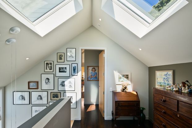 Bright upper-level landing or hallway beneath a steeply sloped ceiling, with two large skylights, white walls, dark timber flooring, framed artwork, and timber furniture. A stair balustrade occupies the foreground, and an open doorway leads to another room. The space has strong natural light and a gallery-like arrangement of artwork.