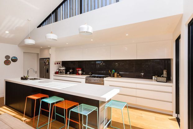 Bright contemporary open-plan kitchen with a large white island, integrated white cabinetry, dark stone-look splashback, stainless-steel appliances, timber flooring, colourful wire-frame bar stools, pendant lighting, and a high-level glazed clerestory creating an airy sense of volume.