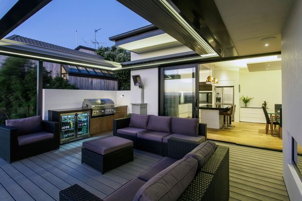 A contemporary covered outdoor entertaining area with dark composite-style decking, extensive roof overhangs, and a large outdoor lounge arrangement. The space includes an outdoor kitchen with stainless-steel barbecue and drinks refrigerator, opening directly into a bright modern kitchen and dining area through wide sliding glass doors. Layered roof forms and clerestory-style glazing create a sheltered indoor-outdoor connection.