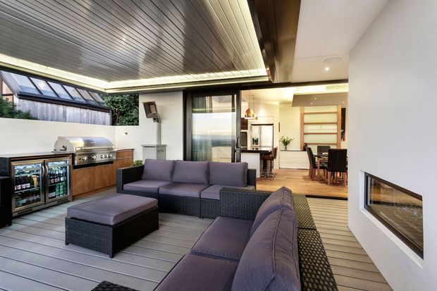 A covered outdoor entertaining area with a built-in barbecue, beverage refrigerators, rattan-style outdoor sofas and ottoman, and a linear gas fireplace set into a white wall. The space opens directly into a bright contemporary kitchen and dining area through large sliding glass doors. A ribbed metal ceiling and grey composite decking define the covered terrace.