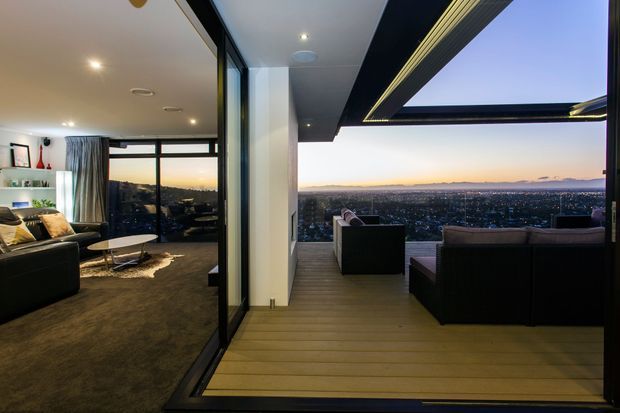 A modern indoor-outdoor living area with a glazed wall opening onto a covered timber-deck terrace. The interior contains dark contemporary seating, a pale rug, recessed ceiling lights, and full-height glazing, while the terrace features outdoor lounge furniture, integrated linear ceiling lighting, and expansive elevated city views at dusk.