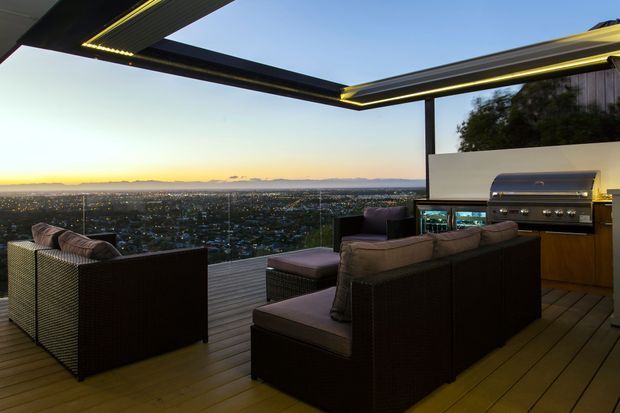 An elevated outdoor entertaining terrace at dusk, featuring dark woven lounge seating, timber decking, a built-in stainless-steel barbecue, and a dramatic cantilevered roof structure with integrated linear lighting. The open edge overlooks a broad cityscape and distant mountains.