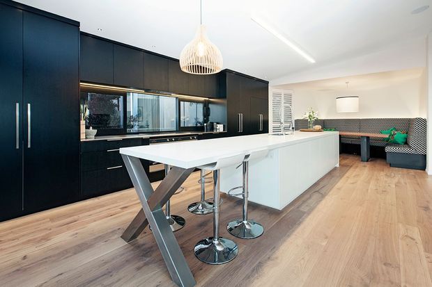 Contemporary open-plan kitchen with a large white island, integrated breakfast bar, dark full-height cabinetry, stainless-steel appliances, timber flooring, and a dining nook with patterned upholstered bench seating. A sculptural pendant light and linear ceiling light add modern decorative accents.