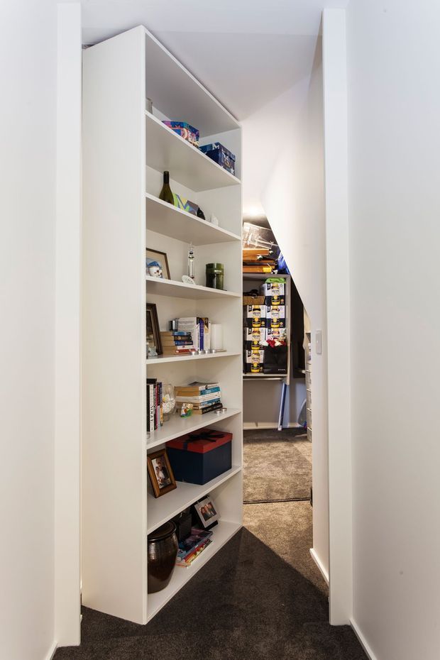 A narrow interior passage with a tall white built-in open shelving unit forming a concealed or pivoting doorway. The shelves display books, framed photographs, decorative objects, bottles, and storage boxes. Dark grey carpet runs through the hallway, with white walls, trim, and ceiling. The opening reveals a compact storage or utility area beyond.