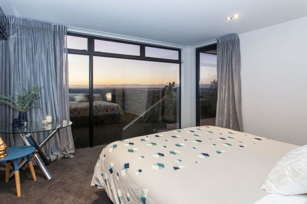 A contemporary bedroom with a large bed facing floor-to-ceiling corner glazing and a broad sunset or landscape view. The room features dark-framed glazed doors/windows, grey curtains, carpeted flooring, white walls, and a patterned neutral bedspread. A small glass side table and decorative chair sit near the glazing.