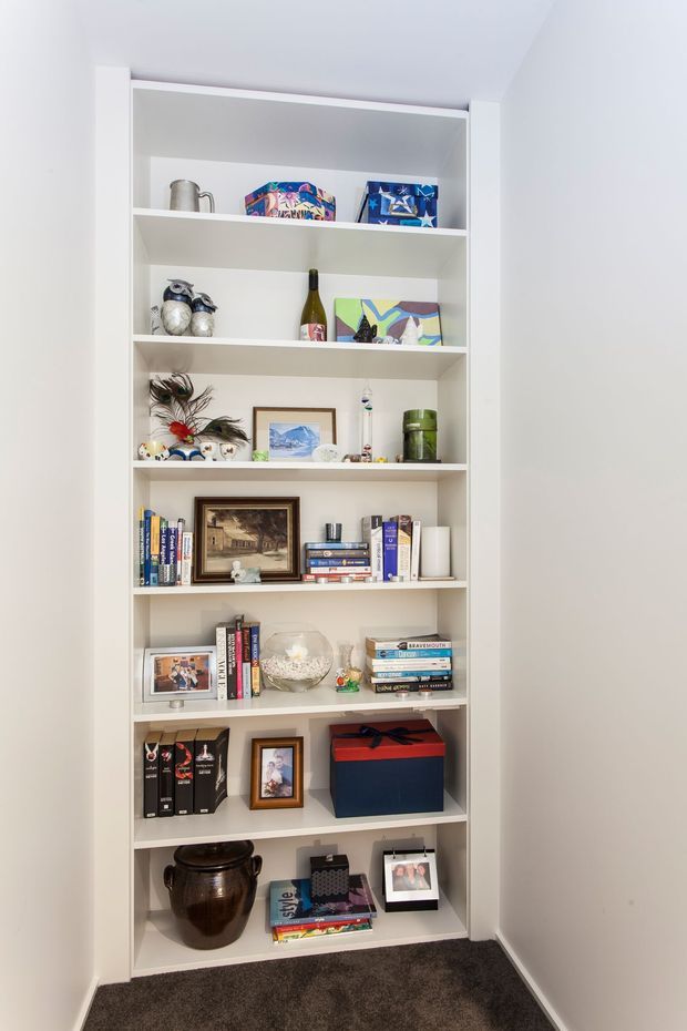A tall built-in white open shelving unit fills a recessed wall niche, displaying books, framed photographs and artwork, ceramics, decorative objects, boxes, and collectibles. The shelving is set against white walls with a dark charcoal carpeted floor, creating a compact display and storage area.