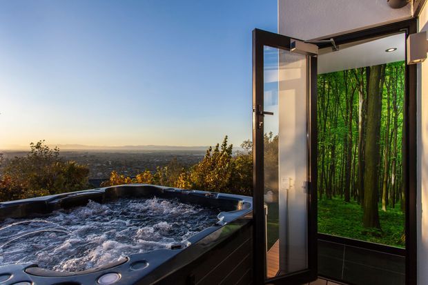 A rooftop or elevated outdoor spa viewed beside an open glass door. The bubbling dark-finished spa occupies the foreground, while the doorway opens onto a compact interior finished with dark wall tiles and a striking forest mural or printed wall surface. Beyond the spa is a broad elevated landscape view at sunset.