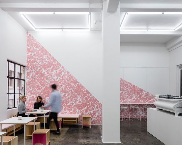 A bright, contemporary commercial interior with a café or communal workspace arrangement. White walls and exposed structural elements are contrasted by a large geometric pink-and-white mural, while small timber tables and stools occupy the polished dark floor. A long counter with a large printer or equipment unit is visible on the right, and black-framed industrial-style windows line the left wall.