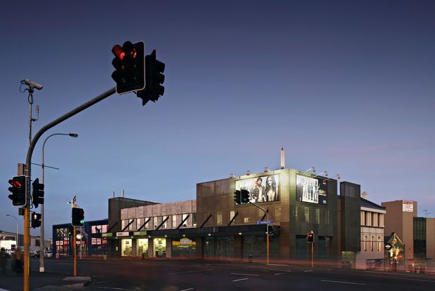 Dusk exterior view of a contemporary multi-storey commercial building on a prominent urban intersection. The facade combines dark brick or masonry panels, large illuminated advertising screens, extensive glazing at ground level, and a flat roofline. Traffic lights, street lighting, road markings, and surrounding retail signage reinforce the urban commercial setting.