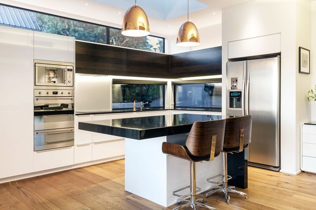 Contemporary kitchen featuring white full-height cabinetry, integrated stainless-steel appliances, a dark timber-toned overhead joinery band, and a large black island benchtop with seating for two. Warm timber flooring, pendant lighting, and expansive windows create a bright, refined space.