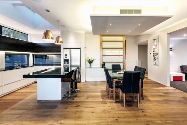 Bright open-plan kitchen and dining area with contemporary white cabinetry, a large dark stone island, integrated stainless-steel appliances, pendant lights, timber flooring, and a glass dining table with dark upholstered chairs. A timber-framed glazed door or window and adjoining living area are visible in the background.