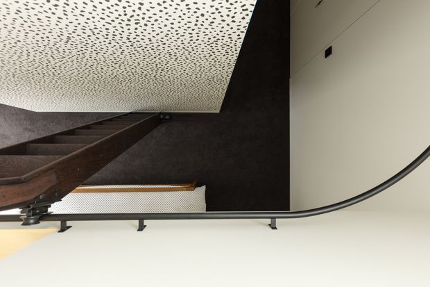 Upside-down view of an interior stairway with a dark timber stair flight, black metal handrail and supports, dark textured wall surfaces, and a distinctive perforated acoustic ceiling. The composition emphasizes architectural detailing and contrast between dark timber/metal and pale ceiling and wall planes.