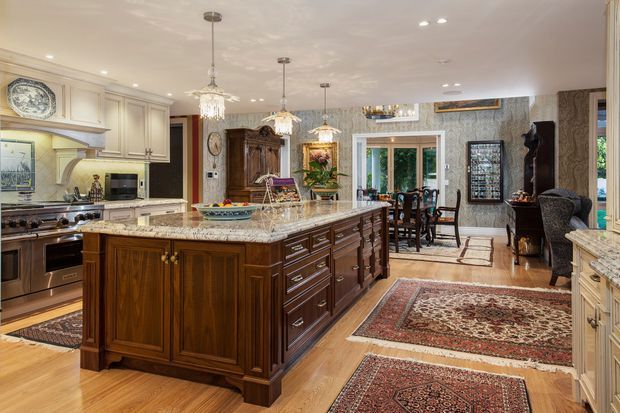 A spacious, richly detailed traditional kitchen with a large dark wood island, ornate cream cabinetry, and a polished light stone benchtop. Crystal pendant lights hang above the island, while patterned wallpaper, framed artwork, decorative objects, Persian-style rugs, and a dining area create a formal, luxurious atmosphere. Timber flooring extends through the open-plan space, with large windows and glazed doors providing natural light.