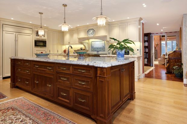 A large traditional kitchen with an ornate dark timber island, pale raised-panel cabinetry, and a light granite or marble benchtop. Decorative pendant lights hang above the island, while patterned rugs, polished timber flooring, framed artwork, and a connected hallway with warm wood finishes reinforce the classic, refined character.