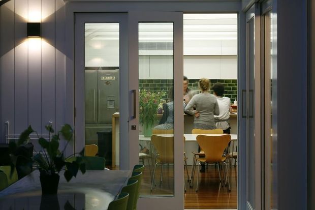 View from a dimly lit adjoining space through glazed grey-framed doors into a bright kitchen and dining area. The kitchen features green glossy subway-tile backsplash, pale cabinetry, stainless-steel appliances, timber flooring, and a dining table with contemporary mustard and green chairs. A dark wall-mounted light and indoor plants frame the foreground.