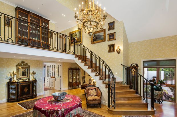 Grand traditional entry hall with a sweeping timber staircase, ornate black wrought-iron balustrades, patterned cream wallpaper, decorative framed artwork, antique display cabinets, and a large gilded chandelier. The room has polished timber flooring, classic furniture, and a mezzanine-level gallery overlooking the foyer.