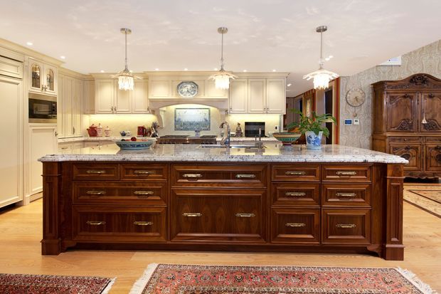 A spacious, traditionally styled kitchen dominated by a large dark timber island with ornate raised-panel cabinetry and polished stone benchtop. Cream painted perimeter cabinetry, integrated appliances, pendant lights, timber flooring, patterned rugs, and decorative furnishings create a warm, highly detailed interior.