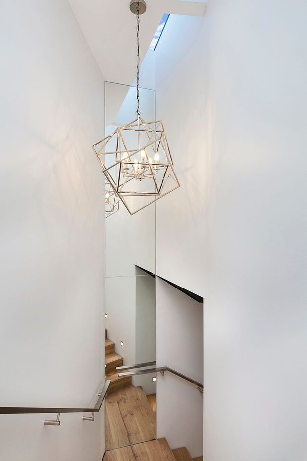 A tall, narrow contemporary stairwell with white walls, timber-finished stair treads and flooring, slim metal handrails, a full-height mirror, and a geometric brass-toned pendant light. A skylight brings natural light into the vertical space.