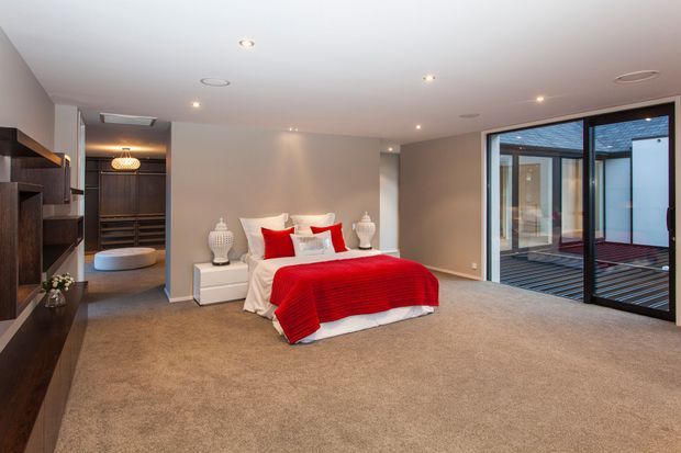 Spacious contemporary bedroom with a low-profile bed dressed in vivid red bedding, dark built-in cabinetry and shelving along the left wall, a walk-in wardrobe or dressing area beyond, and full-height black-framed glazing opening to a small courtyard or terrace. The room has recessed ceiling lighting, neutral painted walls, and plush beige carpet.