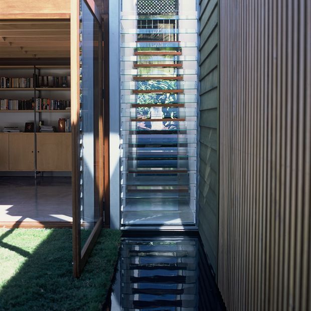 A narrow, light-filled architectural passage or courtyard edge with a full-height glazed louvre screen and reflective dark water feature or polished surface at its base. An open timber-framed glazed door leads into an interior library or living space with built-in timber shelving and cabinetry. The right wall is clad in closely spaced vertical dark timber battens, while the central glazing admits strong daylight and views of planting beyond.