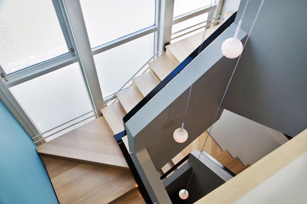 View down a contemporary multi-level stairwell with pale timber stair treads, angular landings, dark painted balustrade panels, and large frosted floor-to-ceiling glazing. Simple spherical pendant lights hang through the stair void, creating a bright, sculptural circulation space.