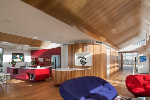A bright, open-plan contemporary interior featuring a vivid red kitchen with island, extensive warm timber flooring and a dramatic timber-clad sloping ceiling. A curved timber feature wall with built-in display artwork separates the kitchen from a lounge area furnished with bold blue and red seating. Large windows and glazing provide abundant natural light and views of an urban setting.