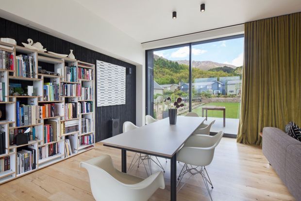 A bright, contemporary dining and reading area with a long white dining table surrounded by sculptural white chairs. A full-height open bookshelf occupies the left wall against dark vertical paneling, while large glazing provides views toward a landscaped suburban setting and mountains. Pale timber flooring, white walls, black ceiling spotlights, and olive-green curtains create a refined, natural-toned interior.