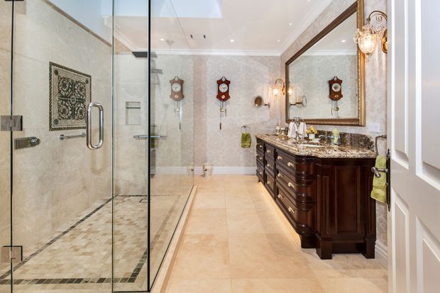 A spacious, traditionally styled bathroom featuring a large frameless glass walk-in shower, pale stone-look wall and floor finishes, ornate dark wood double vanity with a dark patterned stone benchtop, large decorative mirror, crystal wall sconces, and traditional wall clocks. A skylight and recessed ceiling lights provide bright illumination.