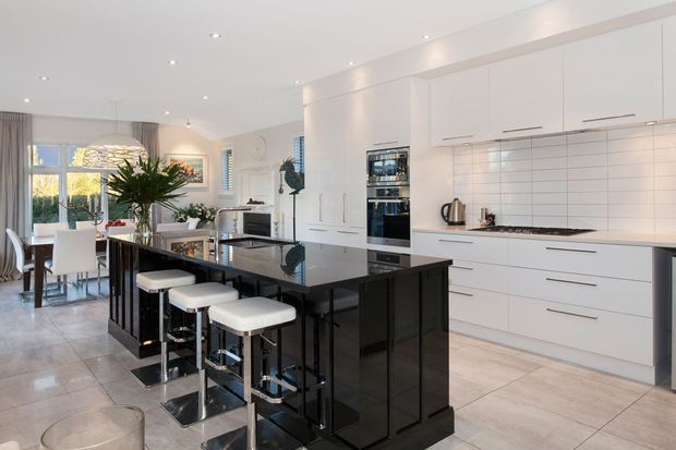 A bright, contemporary open-plan kitchen with extensive white handleless cabinetry, integrated appliances, a glossy black island with seating for four, and a tiled floor. The island separates the kitchen from an adjacent dining area, which is visible alongside large windows and sheer curtains. Recessed ceiling lighting and clean horizontal lines create a polished, high-contrast interior.