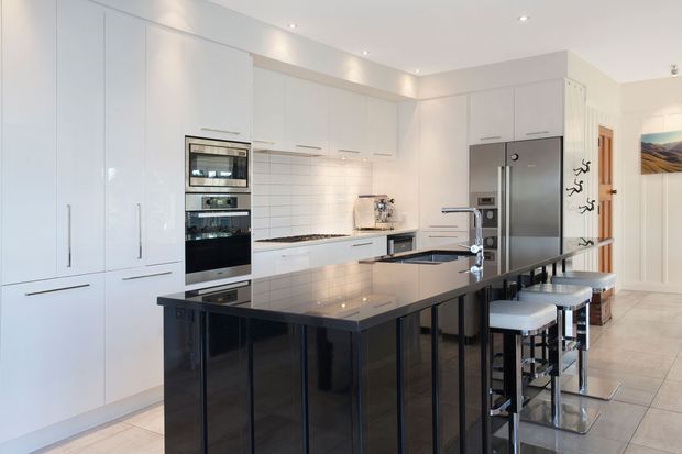 Bright contemporary kitchen with extensive white floor-to-ceiling cabinetry, integrated black appliances, a white tiled splashback, and a large glossy black stone island with bar seating. Recessed ceiling lights and pale flooring create a clean, spacious feel.