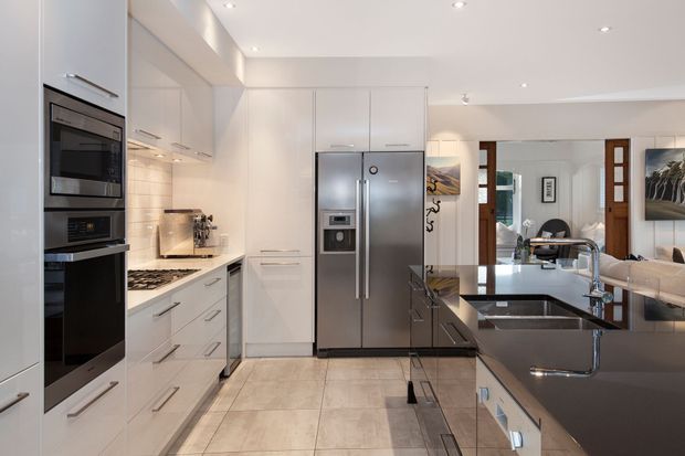 A bright, contemporary kitchen with white full-height cabinetry, integrated stainless-steel ovens, a large stainless-steel French-door refrigerator, pale tiled flooring, and a dark stone island benchtop with an undermount sink and chrome tap. The kitchen opens visually toward a dining and living area through a wide timber-framed opening.