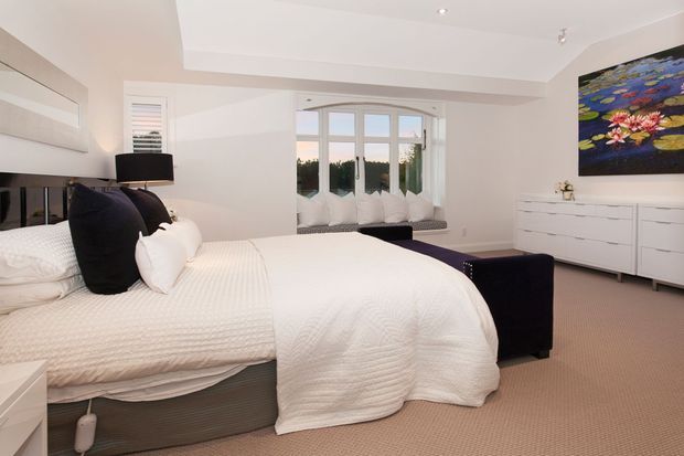 Bright, spacious contemporary bedroom with a large upholstered bed dressed in white textured bedding and contrasting dark pillows. The room has pale walls, beige carpet, a white dresser, a black bedside lamp, a dark upholstered bench or ottoman, broad windows with white plantation shutters, recessed ceiling lighting, and a colourful floral artwork above the dresser.