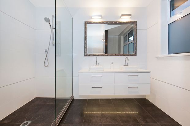 A bright, contemporary bathroom with a frameless glass walk-in shower, dual wall-mounted vanity basins, white built-in cabinetry, a large decorative framed mirror, white wall surfaces, and dark tiled flooring. A window provides additional natural light.