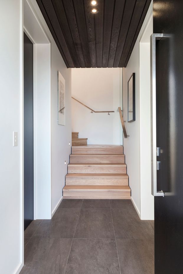 A contemporary stairway viewed from an entry hall, with broad light timber treads, dark tiled flooring, white walls, slim metal handrails, and a striking dark timber-lined ceiling with recessed downlights. Black-framed artwork and a dark door add contrast.