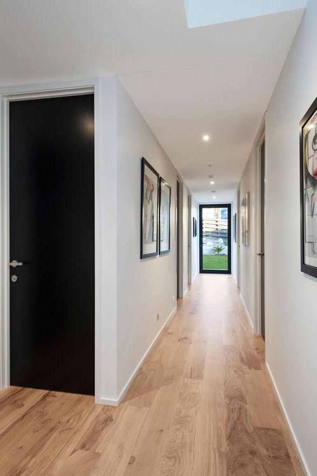 A bright, narrow residential hallway with light timber flooring, white walls and ceiling, recessed downlights, framed artwork, and several white interior doors. A matte black door stands open on the left, while a glazed door at the far end provides a view toward an outdoor area and lawn.