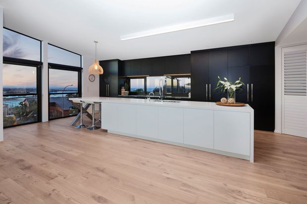 Bright contemporary kitchen with a large white island, black full-height cabinetry, timber flooring, black-framed floor-to-ceiling glazing, and a small dining nook. The glazing overlooks a coastal or waterfront setting, while the interior is clean-lined and open plan.