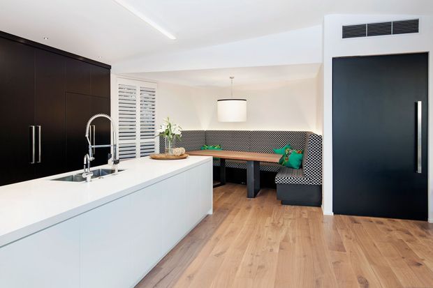 A bright, contemporary open-plan kitchen and dining nook. The kitchen features a large white island with an integrated sink and spring-coil faucet, white cabinetry, and a contrasting wall of dark cabinetry. Beyond the island is a built-in dining booth upholstered in black-and-white patterned fabric, paired with a dark dining table and pendant light. Light timber flooring, white walls, plantation shutters, and a black internal door complete the space.