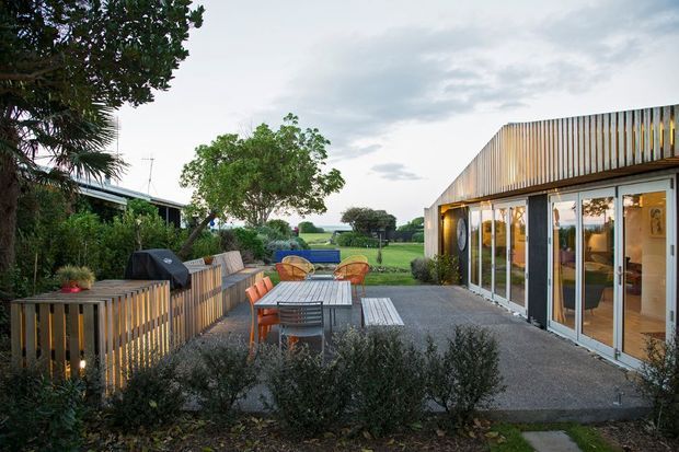 A landscaped outdoor entertaining area beside a contemporary single-storey building. The paved terrace includes a long timber dining table with bench seating and colourful chairs, bordered by timber slat fencing, raised planting beds and shrubs. Full-height glazed sliding doors open into a warmly lit interior, while a low sloping metal-clad roof and vertical timber screening define the building facade. Rural greenery is visible beyond the property.