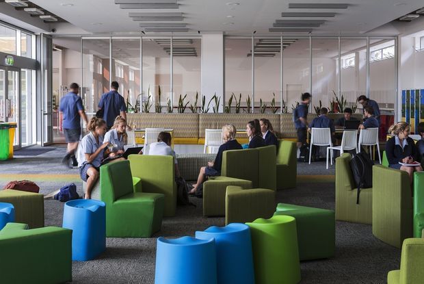 A bright, contemporary communal interior resembling a school or institutional breakout and dining area. The open-plan space contains green upholstered lounge chairs and ottomans, blue sculptural side tables, booth seating with planters, and groups of people working or socialising. Glazed partitions divide adjacent spaces, while large windows and extensive linear acoustic ceiling panels provide a light, functional environment.